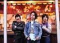 The three men of Electric Eel Shock are standing in front of a Japanese eatery, identifiable by the banner with the word "おでん" (Oden) on it. The frontman stands in the centre, wears a denim jacket and has his hands in his pockets, while his companions on either side display different postures with one having his arms crossed and the other pointing a finger towards probably the eatery. © Photo by Yuki Kuroyanagi