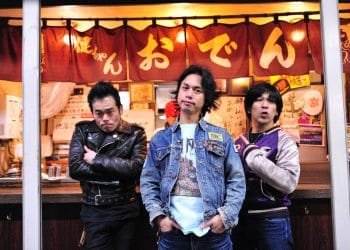 The three men of Electric Eel Shock are standing in front of a Japanese eatery, identifiable by the banner with the word "おでん" (Oden) on it. The frontman stands in the centre, wears a denim jacket and has his hands in his pockets, while his companions on either side display different postures with one having his arms crossed and the other pointing a finger towards probably the eatery. © Photo by Yuki Kuroyanagi