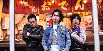 The three men of Electric Eel Shock are standing in front of a Japanese eatery, identifiable by the banner with the word "おでん" (Oden) on it. The frontman stands in the centre, wears a denim jacket and has his hands in his pockets, while his companions on either side display different postures with one having his arms crossed and the other pointing a finger towards probably the eatery. © Photo by Yuki Kuroyanagi
