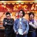 The three men of Electric Eel Shock are standing in front of a Japanese eatery, identifiable by the banner with the word "おでん" (Oden) on it. The frontman stands in the centre, wears a denim jacket and has his hands in his pockets, while his companions on either side display different postures with one having his arms crossed and the other pointing a finger towards probably the eatery. © Photo by Yuki Kuroyanagi