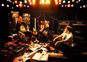 Photo of the three band members of the alternative heavy rock band ZEROSHIKI. They are all sitting down on a live stage, surrounded by instruments and amplifiers. They are sitting in a circle, as if they have a band meeting and have food and drinks (sushi, sake, Pocky, takoyaki) in front of them, but one of the members also has a laptop in front of him. All are looking at the camera with a smile, two of them are holding their arms halfway up, making it seem that the photo was taken during a joyful moment.
