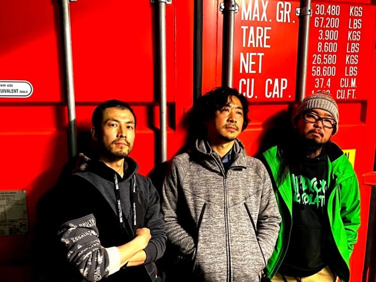 The three members of ZEROSHIKI standing against a red shipping container © ZEROSHIKI