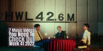 Still from Bob Paulsen's music video for "Doggs". The three band members of the rock band are sitting at a table at a seemingly outside location, waiting for their dinner.