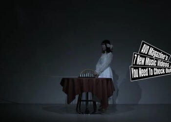 Still from the music video for "毒々(Dokudoku)" by HAIMEN WORKS. A girl dressed in white clothing is blindfolded and stands next to a chair. In front of her is a table with a game of chess.