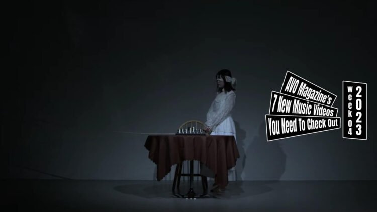 Still from the music video for "毒々(Dokudoku)" by HAIMEN WORKS. A girl dressed in white clothing is blindfolded and stands next to a chair. In front of her is a table with a game of chess.