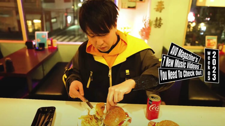 Still from Gasoline Coke's music video for the song "バーガー (Burger)". A man - wearing a black and yellow jacket - is at a well-lit restaurant cutting his hamburger in pieces before decided to just eat it with his hands.