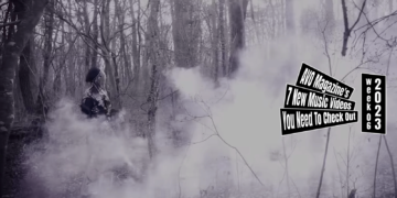 Still from HOWL's music video for "アンダーテイカー (Undertaker)". Vocalist Mayoi is walking through a partly misty forest, while dressed in dark clothing and wearing a beret. The colouring of this still is monochrome.