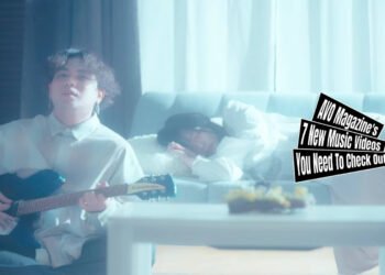Still from the music video "901" by 海とクレヨン (Umi to Crayon). The vocalist and guitarist of 海とクレヨン (Umi to Crayon) plays on his guitar while sitting on the ground, leaning against a couch where a girl is sleeping on. The room they are in is lightened up and both persons are wearing light clothing making the situation almost angelic.