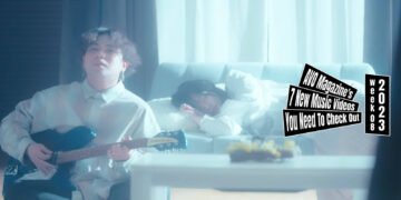 Still from the music video "901" by 海とクレヨン (Umi to Crayon). The vocalist and guitarist of 海とクレヨン (Umi to Crayon) plays on his guitar while sitting on the ground, leaning against a couch where a girl is sleeping on. The room they are in is lightened up and both persons are wearing light clothing making the situation almost angelic.