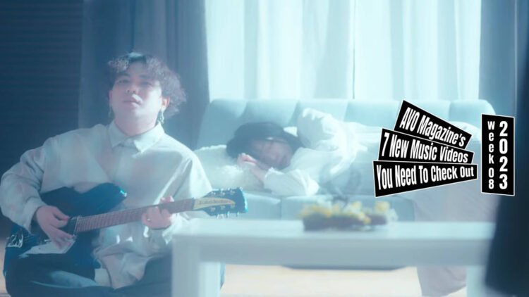 Still from the music video "901" by 海とクレヨン (Umi to Crayon). The vocalist and guitarist of 海とクレヨン (Umi to Crayon) plays on his guitar while sitting on the ground, leaning against a couch where a girl is sleeping on. The room they are in is lightened up and both persons are wearing light clothing making the situation almost angelic.