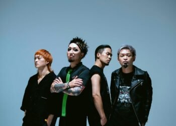 The four band members of reggae punk band SiM, dressed in mostly dark clothing are standing next to each other while looking in the camera with serious faces.