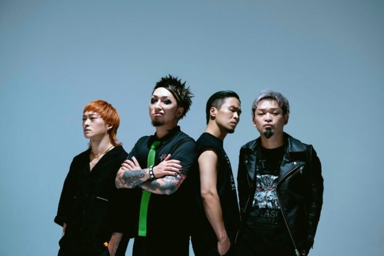 The four band members of reggae punk band SiM, dressed in mostly dark clothing are standing next to each other while looking in the camera with serious faces.