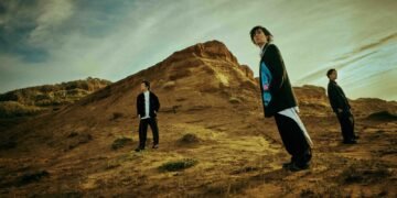 The members of RADWIMPS are standing on what seams a sandy hill near a beach. They all wear mostly dark clothing and only the vocalist and guitarist looks in the direction of the camera.