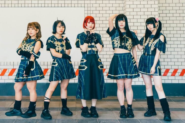 The five members of idol group DEMO, they are standing next to each other while posing and looking to the person who took the photo. They are dressed in feminine clothing with blue and green stripes, black patches and golden coloured embellishments.