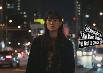 A still from the music video for "3" by the band Sandi gray eyes. The vocalist and guitarist is standing near a busy street during nighttime, vehicles are driving past her and she is surrounded by lights while she is singing a song.