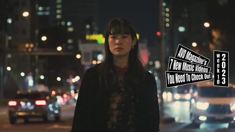 A still from the music video for "3" by the band Sandi gray eyes. The vocalist and guitarist is standing near a busy street during nighttime, vehicles are driving past her and she is surrounded by lights while she is singing a song.