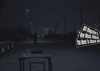 Still from polly's music video for the song "Kikoeru". Polly's vocalist and guitarist sat at a school table during the music video and in this still, the table and chair is placed at a road in a silent village, during the dark hours of the day.