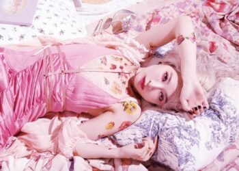 Nana Kitade is dressed in baby pink clothing and posing while seemingly lying on pillows and blankets.