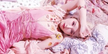 Nana Kitade is dressed in baby pink clothing and posing while seemingly lying on pillows and blankets.