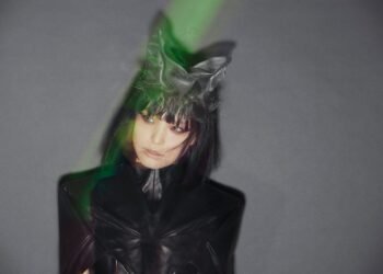 Image of RIS of solo project RIS-707 dressed in fully black and on her head what looks like cat ears, behind her is a grey background. She does not look at the camera but looks on the side.