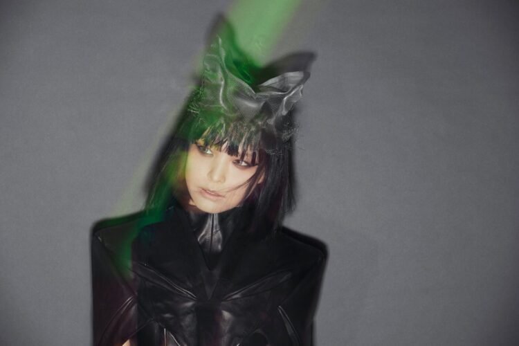Image of RIS of solo project RIS-707 dressed in fully black and on her head what looks like cat ears, behind her is a grey background. She does not look at the camera but looks on the side.