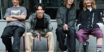 The four members of ONE OK ROCK are sitting on chairs, the vocalist of the band, TAKA, is leans forward and smiles into the camera. The other three members are also smiling but are look away from the camera.