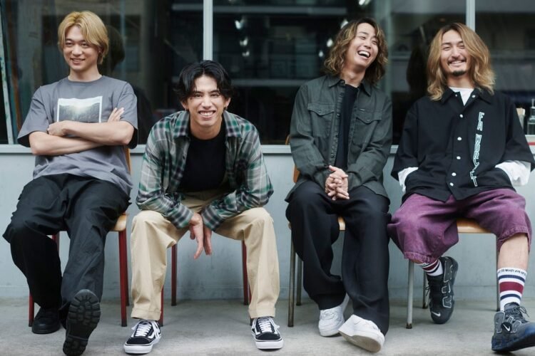 The four members of ONE OK ROCK are sitting on chairs, the vocalist of the band, TAKA, is leans forward and smiles into the camera. The other three members are also smiling but are look away from the camera.