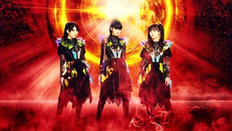 The three members of BABYMETAL are posing while being surrounded of what looks like fire.