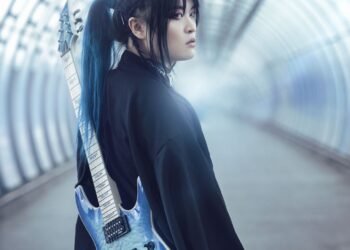 Band leader Kai is dressed in black clothing and carries a soft blue-coloured electric guitar on the back. Kai seems to be walking on an empty train station or in an empty tunnel and looks behind him.