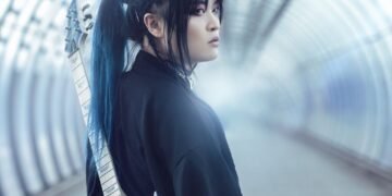 Band leader Kai is dressed in black clothing and carries a soft blue-coloured electric guitar on the back. Kai seems to be walking on an empty train station or in an empty tunnel and looks behind him.