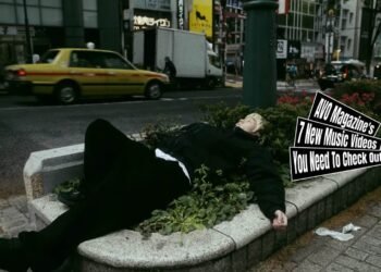 Still from the music video "Stressed Town" by the band LOBSTER. One of the band members of LOBSTER, the vocalist, is dressed in dark clothes and bleached hair lies in a bush near a busy street. The music video starts with him sleeping in that same location and the video further shows how he ends up there.