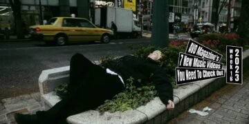 Still from the music video "Stressed Town" by the band LOBSTER. One of the band members of LOBSTER, the vocalist, is dressed in dark clothes and bleached hair lies in a bush near a busy street. The music video starts with him sleeping in that same location and the video further shows how he ends up there.