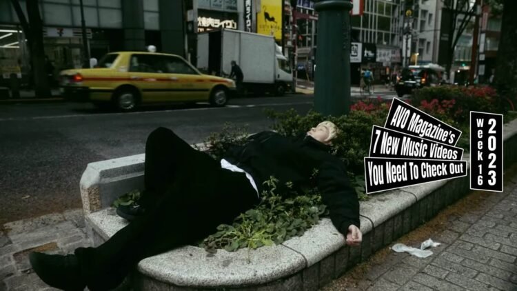 Still from the music video "Stressed Town" by the band LOBSTER. One of the band members of LOBSTER, the vocalist, is dressed in dark clothes and bleached hair lies in a bush near a busy street. The music video starts with him sleeping in that same location and the video further shows how he ends up there.