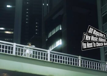 Still from AMUSEMENT LAGER's music video for the song "White". A girl is standing listening to music through her headphones and looking out from a bridge to enjoy the view during nighttime. This shot was shot from the ground upwards. This still has been mirrored.