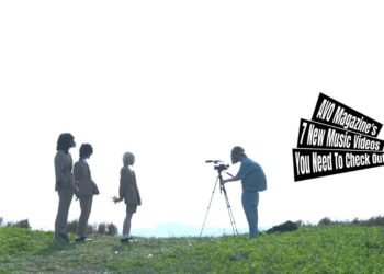 Still from the music video by セグウェイライダー (Segway Rider) for the song "ライフスタイル (Lifestyle)". The three members of Segway Rider are suited up and are standing on the grass in front of a camera man with a mask and the camera. The weather is sunny in this still.
