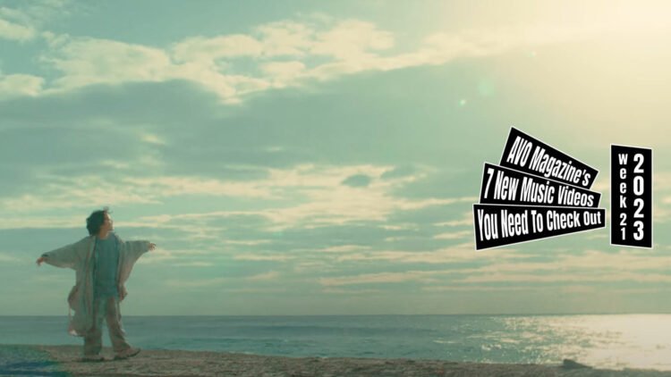 Still from Awkmiu's music video for the song "avalanche". The vocalis, dressed in a blueish coloured clothing, blue shirt, pants and an oversized cardigan or jacket (the lighting/editing makes it seem that the clothing is different in colour), is on the beach, dancing, spinning around and singing.