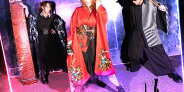 The three members of AKARA are standing next to each other, in front is the vocalist and a bit behind her the musicians with their instruments.