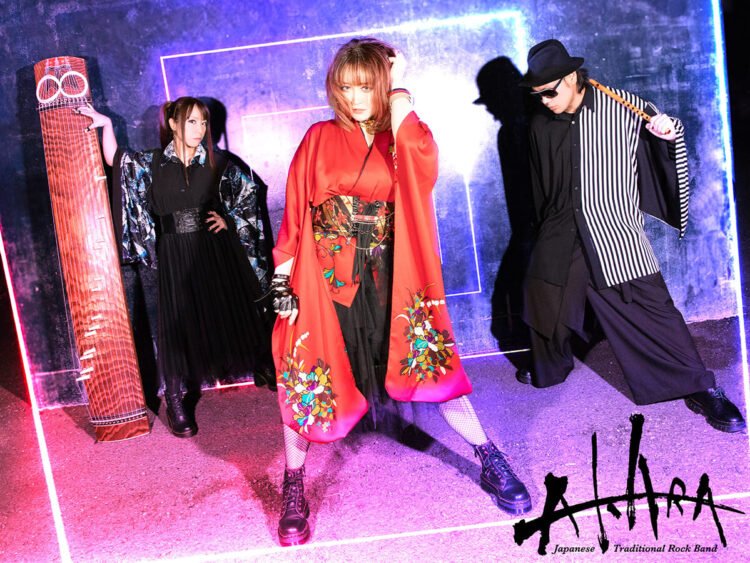 The three members of AKARA are standing next to each other, in front is the vocalist and a bit behind her the musicians with their instruments.
