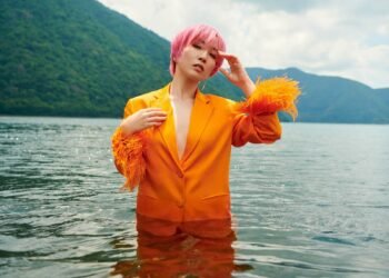 Nao Yoshioka with pink hair and orange suit standing (and posing) in the middle of a lake.