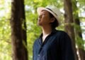 Yutaro Ogida stands in front of a couple of trees and looks away from the camera. He is wearing a dark blue shirt and a fedora.