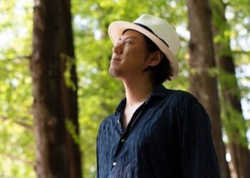 Yutaro Ogida stands in front of a couple of trees and looks away from the camera. He is wearing a dark blue shirt and a fedora.