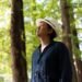 Yutaro Ogida stands in front of a couple of trees and looks away from the camera. He is wearing a dark blue shirt and a fedora.