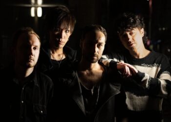 The four current members of ASK I FALL (in 2023) posing a dark area with limited light sources allowing the faces of the band members to be visible on one side.