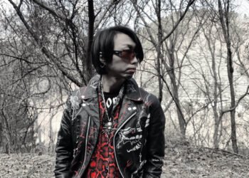 DEATHROLL's Kazu in a leather jacket and sunglasses stands by a pair of leafless trees.