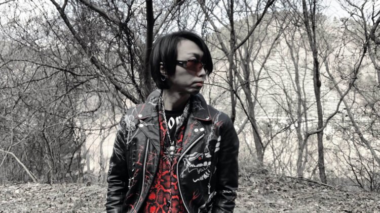 DEATHROLL's Kazu in a leather jacket and sunglasses stands by a pair of leafless trees.