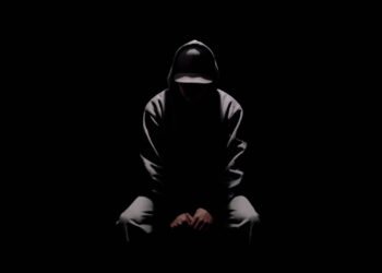 Ryo Kinoshita sits on a stool while surrounded by darkness and only one lamp shines light on him. His face is also not visible in the picture, giving it a mysterious appearance.