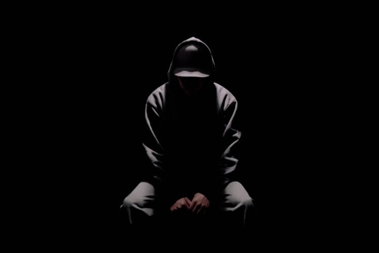 Ryo Kinoshita sits on a stool while surrounded by darkness and only one lamp shines light on him. His face is also not visible in the picture, giving it a mysterious appearance.
