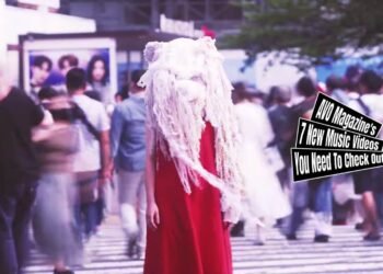 Still from Kako Fujita's music video for the song "怪物 (Kaibutsu)". A woman with a special costume - made by Emi Fukumoto - consisting of a red dress and a white-coloured artsy headpiece stands in the middle of a busy pedestrian crossing.