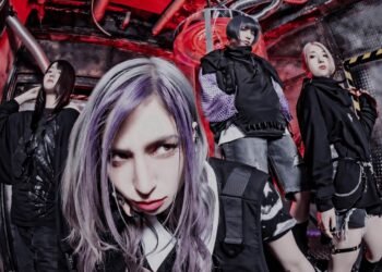 This image shows a group of women that are part of the Japanese band Little Lilith in various costumes, gathered together for a photo. The majority of the group wears black clothing, with some variations in style and colour. The photo seems to be taken in an industrial space.