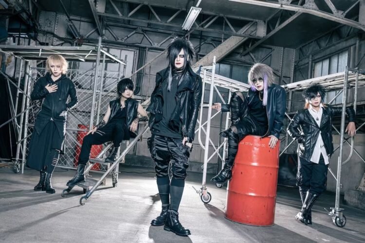 The photo shows a group of five people in black clothing standing inside what seems to be an industrial space. All five musicians appear to be looking off into different directions while maintaining their composure despite being surrounded by such an industrial setting.